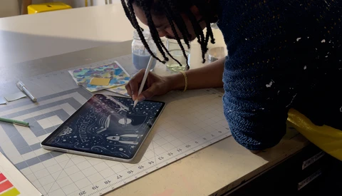 Person drawing on a dark blue patterned surface with a pen while seated at a white desk with a ruler and other materials nearby.