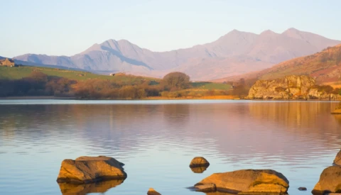 Mountain lake at golden hour with foreground rocks and tree-lined shoreline under clear sky.