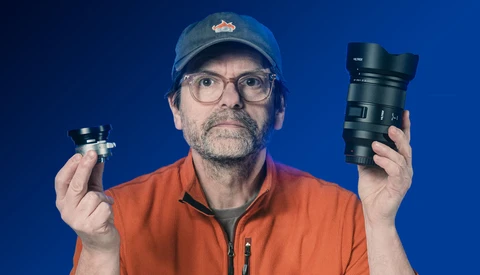 Photographer holding a compact prime lens in one hand and a larger telephoto lens in the other against a blue background.