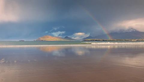 Rainbow stretching across a bay toward mountains reflected in wet sand at golden hour.