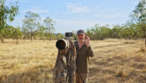 Wildlife photographer holding a telephoto lens in an open woodland savanna landscape.