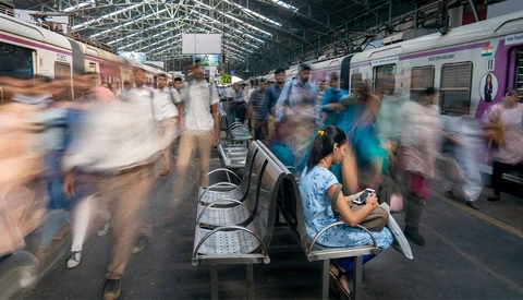 Woman sitting on a bench at a train station, surrounded by blurred commuters and purple trains.