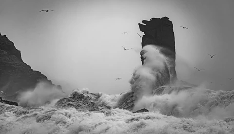 Dramatic black and white seascape with towering rock formation engulfed in crashing waves and mist.