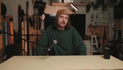 Man speaking into microphone at table in workshop with tools and lighting equipment in background.