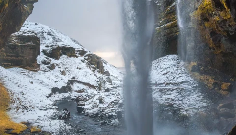 Tall waterfall cascading down snow-covered rocky cliffs with golden lichen visible on the stone.
