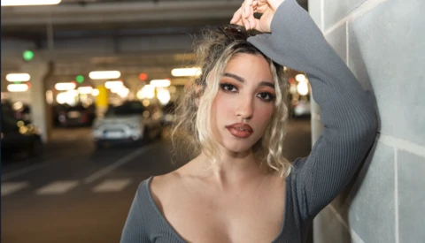 Woman with blonde hair posing against a white wall in a parking garage setting.