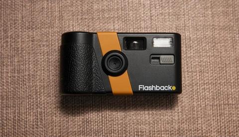 Flashback vintage film camera with black body and orange stripe on beige fabric background.