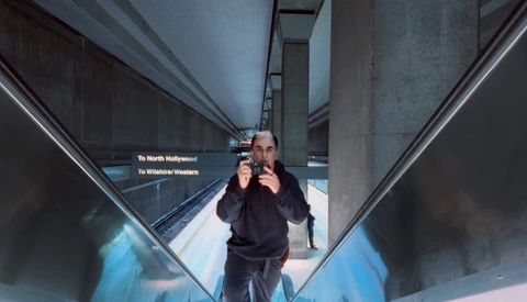 Man in black hoodie standing in subway station corridor with directional signs visible overhead.