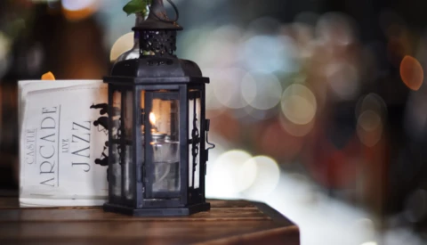 Black metal lantern with candle flame beside a white candle container, shot with shallow depth of field and warm bokeh background.