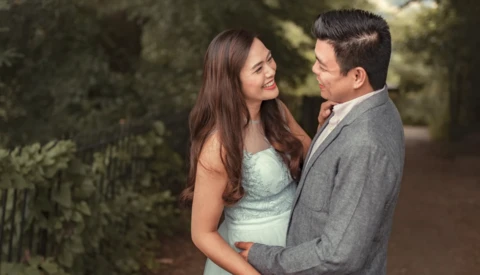 Couple embracing outdoors among green foliage in soft natural light.