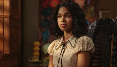 Woman with long dark curly hair wearing a cream-colored blouse with puff sleeves and tie neckline, looking off-camera in an interior setting.