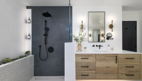 Modern bathroom interior with dark tile shower enclosure, wooden vanity, and brass-accented mirrors and lighting fixtures.