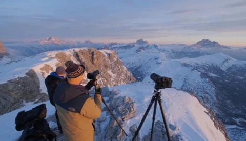 Photographer on snowy alpine ridge with camera on tripod overlooking snow-covered peaks at golden hour.