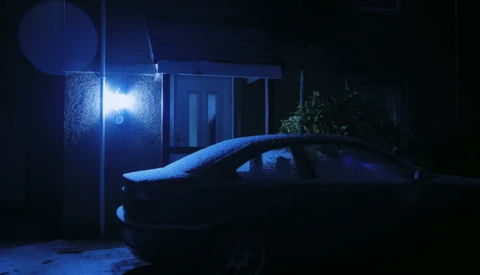 Parked sedan covered in snow beneath a blue-tinted streetlight at night.