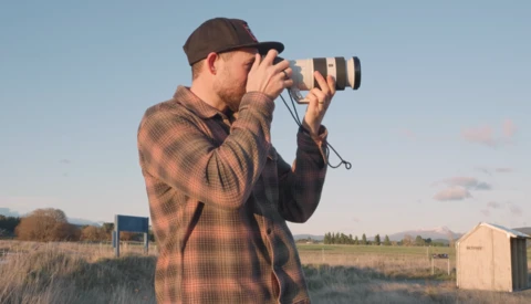 Photographer in plaid shirt and cap using telephoto lens outdoors at golden hour.