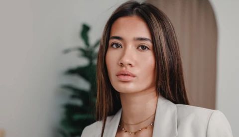 Woman in cream blazer with long dark hair against neutral interior background.