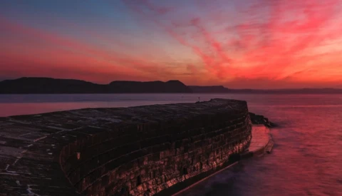 Rocky headland silhouetted against a vibrant crimson sunset reflected in still water.