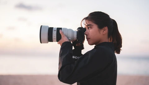 Photographer with long dark hair using a telephoto lens during golden hour at a coastal location.