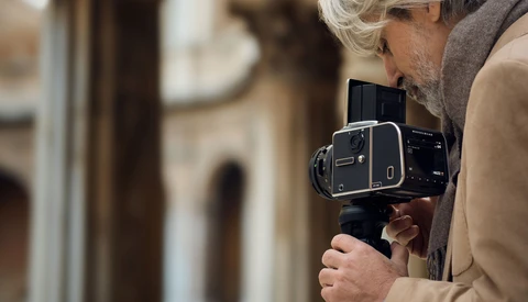 Photographer with white hair holding a large format film camera to their eye in an urban courtyard setting.