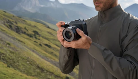 Photographer holding a mirrorless camera while standing in a mountainous landscape with green slopes.