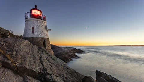 White stone lighthouse with red top perched on rocky coastal outcrop at sunset, its beacon illuminated against twilight sky.