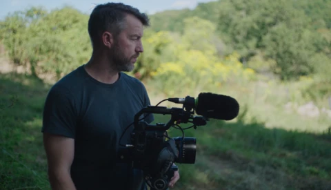 Videographer holding a professional cinema camera with shotgun microphone in an outdoor natural setting.