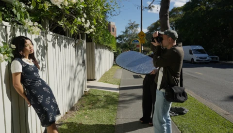 Photographer holding large reflector while shooting portrait of woman against white fence in natural daylight.