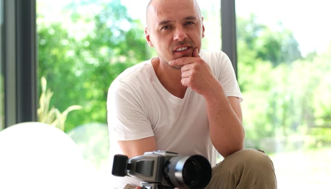 Male photographer holding a professional video camera while posing indoors near large windows with natural landscape visible outside.