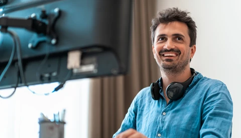 Man in denim shirt smiling at camera in a studio setup with monitor and headphones.