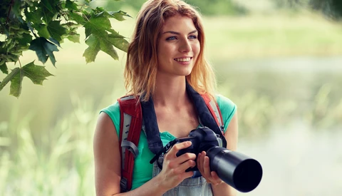 Woman holding binoculars outdoors near tree branches and blurred water background.
