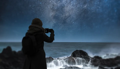 Silhouetted figure photographing the Milky Way above turbulent ocean waves at night.