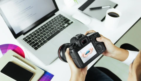 Photographer's hands holding a DSLR camera displaying an image on its rear LCD screen, positioned above a desk workspace.