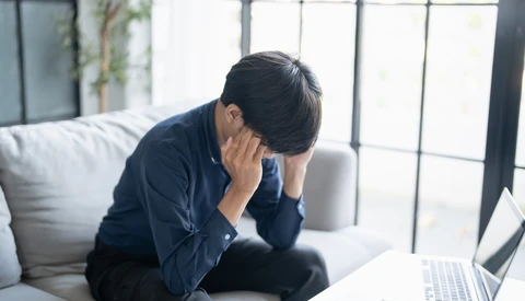 Man in dark blue shirt holding his head while sitting at laptop, expressing stress or fatigue.
