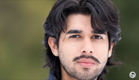 Portrait of a man with shoulder-length dark hair and facial hair against a soft green background.