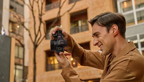Photographer holding a vintage film camera up to his face, smiling while framing a shot in an urban setting.