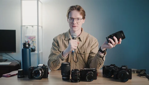 Man holding a camera lens while discussing photography gear displayed on a table.