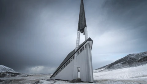 Modern white chapel with tall spire tilted dramatically in snowy Arctic landscape under stormy sky.
