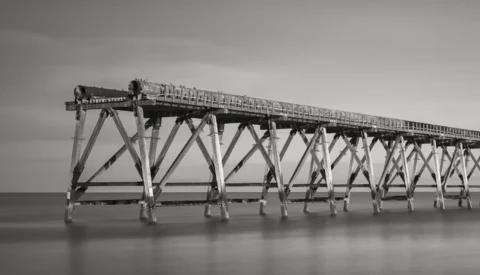 Long wooden pier with repetitive X-braced support structure extending into calm water under overcast sky.