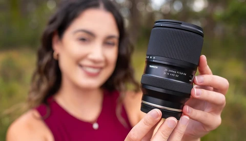 Woman holding a telephoto lens toward the camera in an outdoor garden setting.