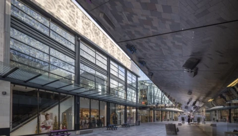 Modern transit station with expansive skylights, retail storefronts, and geometric ceiling architecture.