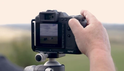 Photographer holding a medium format camera on a tripod, viewing the outdoor landscape through the LCD screen.