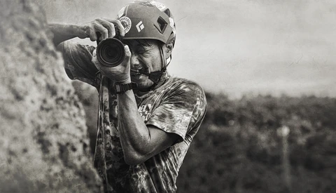 Climber in helmet peers through camera lens while secured to rock wall.