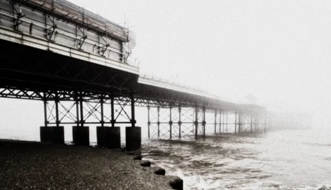 Pier structure receding into thick fog with dark metal framework and support pillars extending into turbulent water.