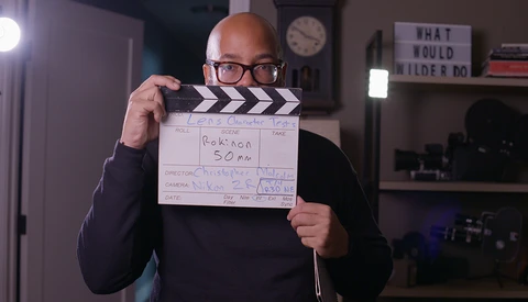 Bald man wearing glasses holds a film clapperboard marked "GARCON SOM" in a home studio setting.