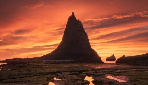 Dramatic silhouette of a tall sea stack against a vibrant orange and yellow sunset sky with reflection in wet tidal flats.