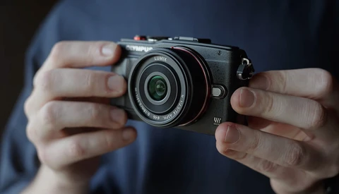 Hands holding a compact mirrorless camera with a fixed prime lens against a blue shirt.