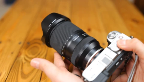 Hands holding a mirrorless camera with a telephoto lens mounted on a wooden surface.