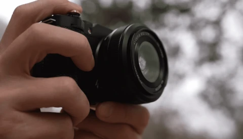 Hands holding a compact mirrorless camera with a standard zoom lens, photographing outdoors.