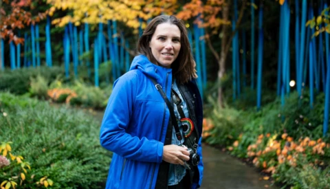 Woman holding a camera wearing a bright blue jacket in an autumn garden setting.