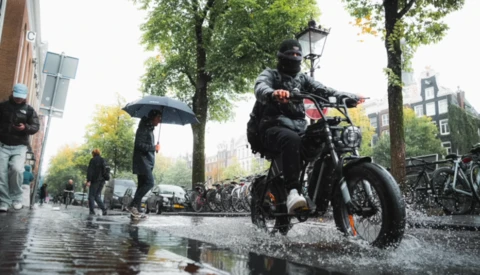 Man riding a motorcycle through a wet urban street during rain, splashing through water.